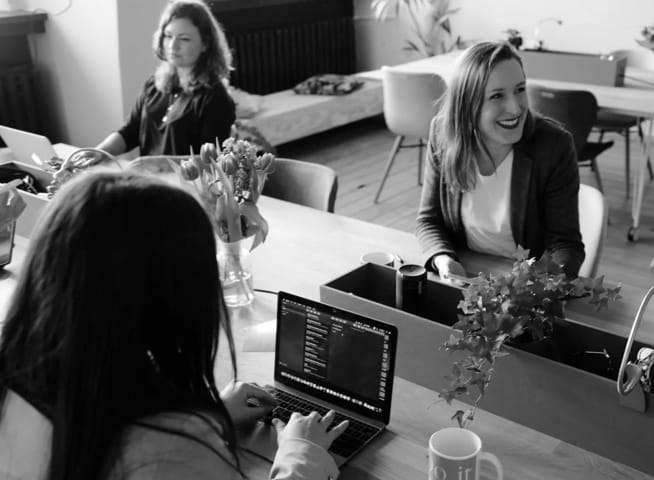 Women sitting at a table chatting and typing on laptops.